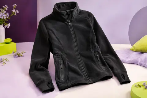 Veste polaire noire à col haut, manches à trous pour les pouces et poches zippées posée sur un fond de studio lilas.