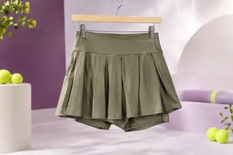 Jupe-short de tennis kaki plissée à taille haute, posée sur un fond de studio lilas et ivoire.